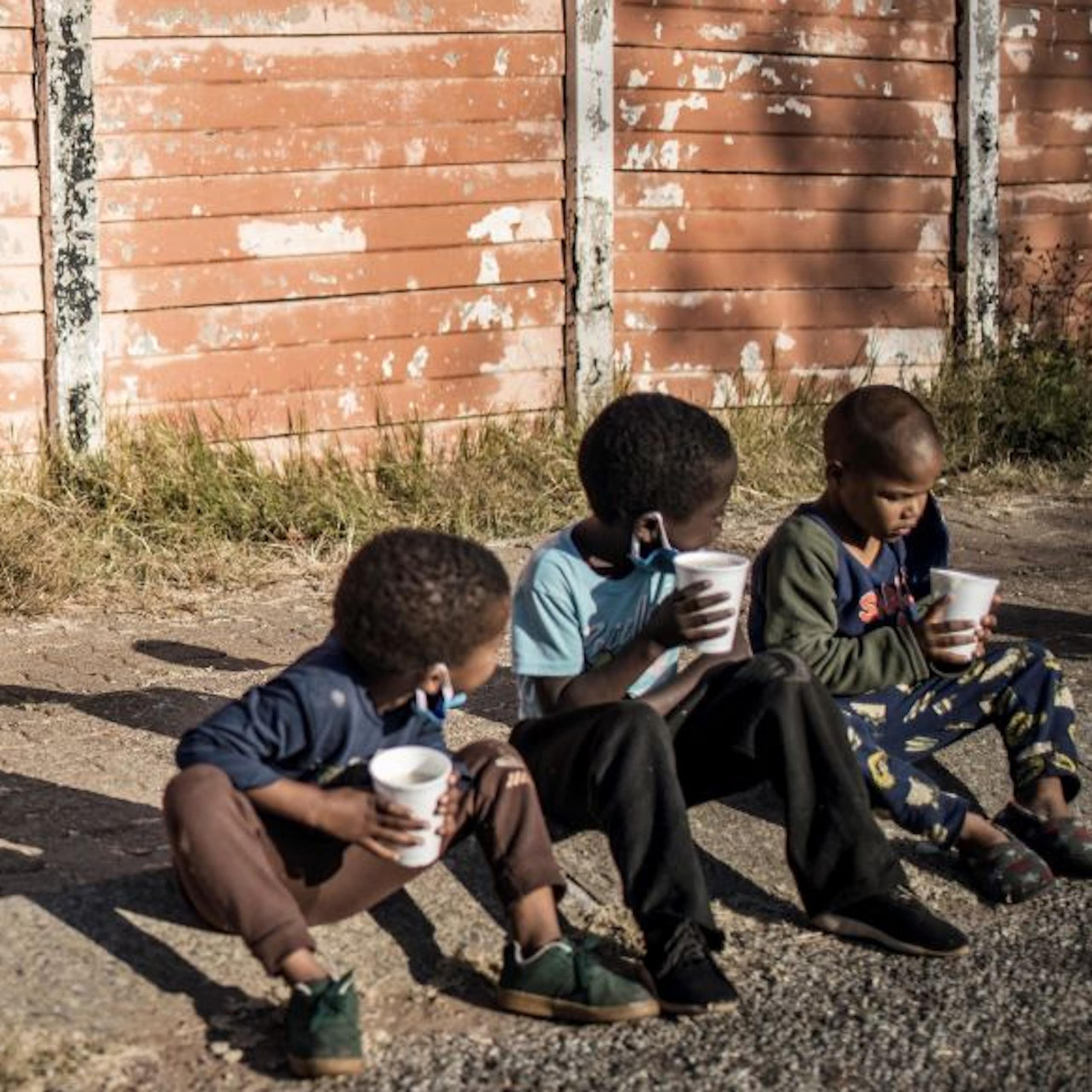 Three young children sit on a pavement, holding disposable cups, while a dog sniffs the ground behind them