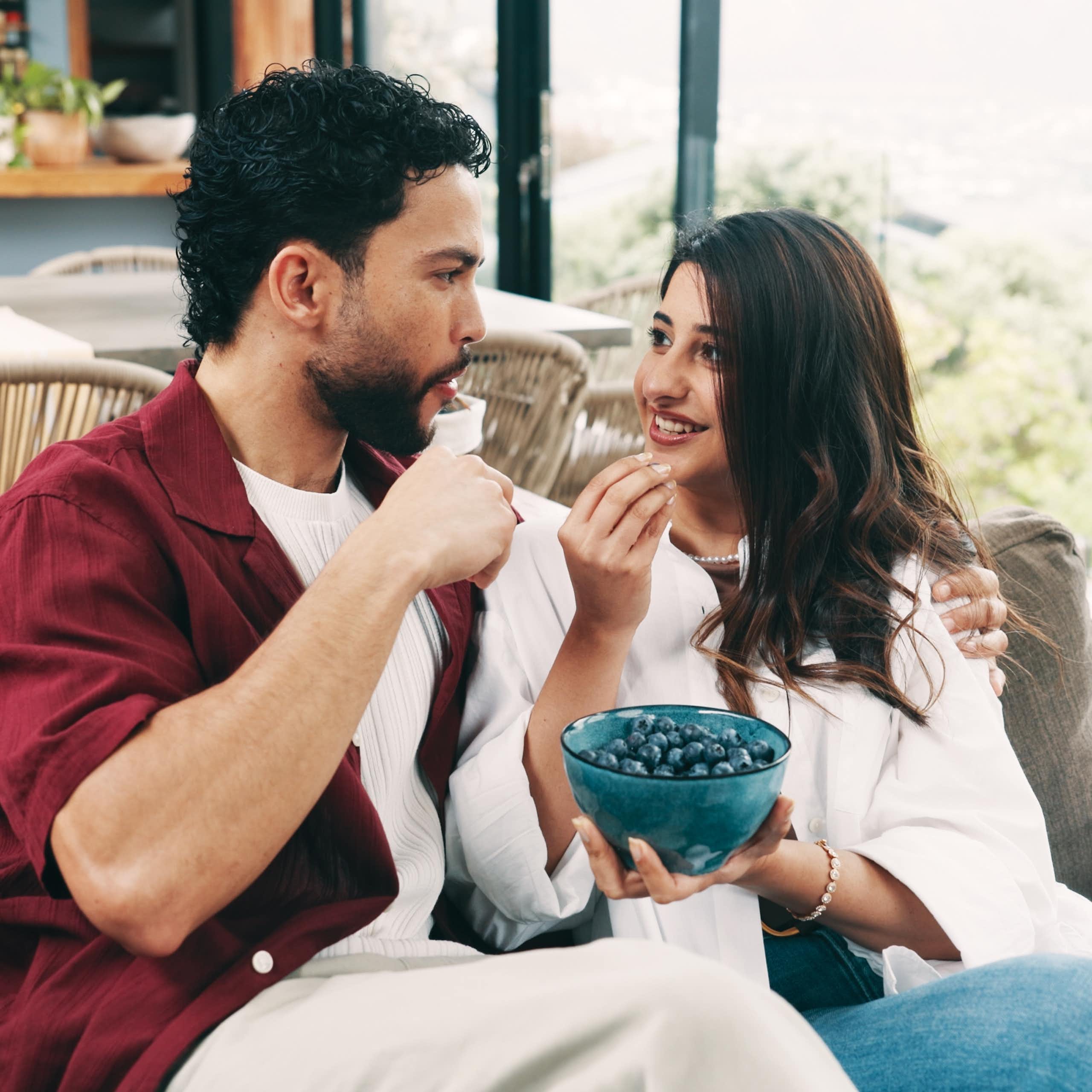A couple sitting next to each other on a sofa, sharing a bowl of blueberries.