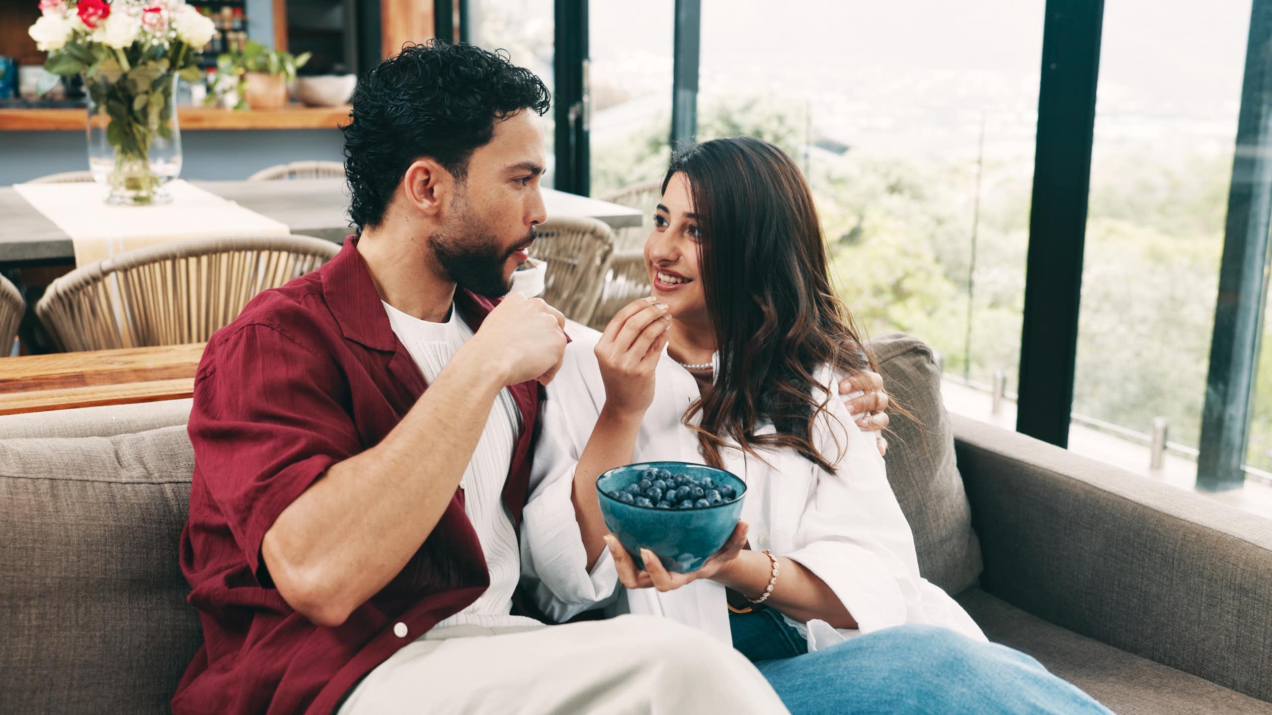A couple sitting next to each other on a sofa, sharing a bowl of blueberries.