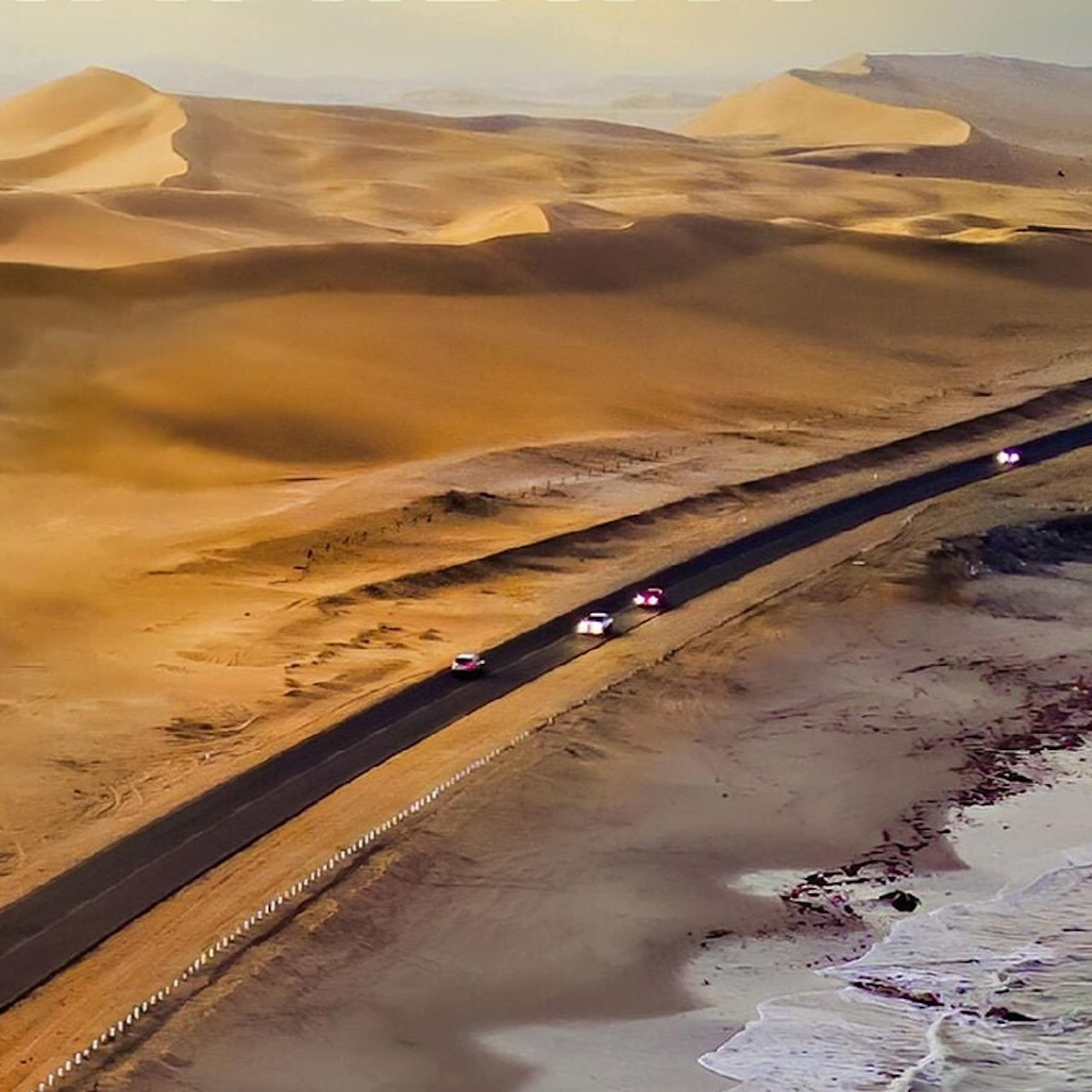 A coastline with a road containing cars and vast sand dunes on the other side of it.