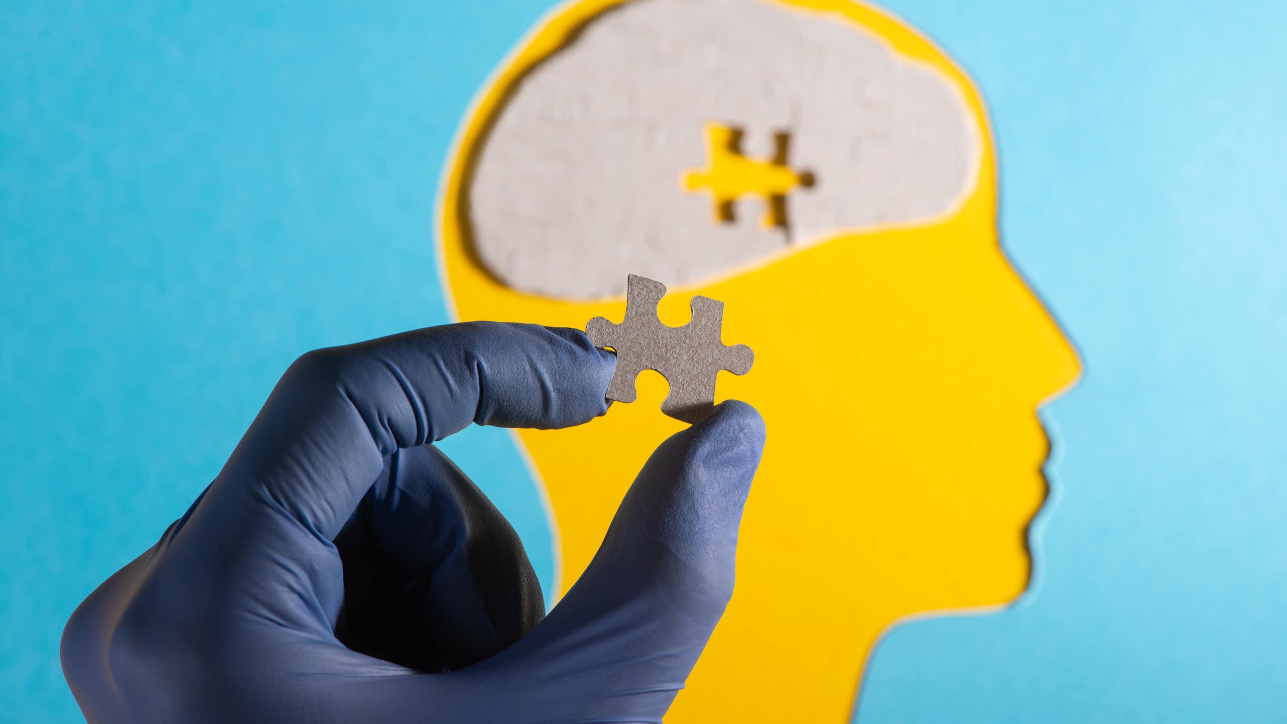 A cutout of a head in side-profile. In the centre of the head's brain, it is missing a puzzle piece. A person's hand holds that missing piece between their thumb and forefinger.