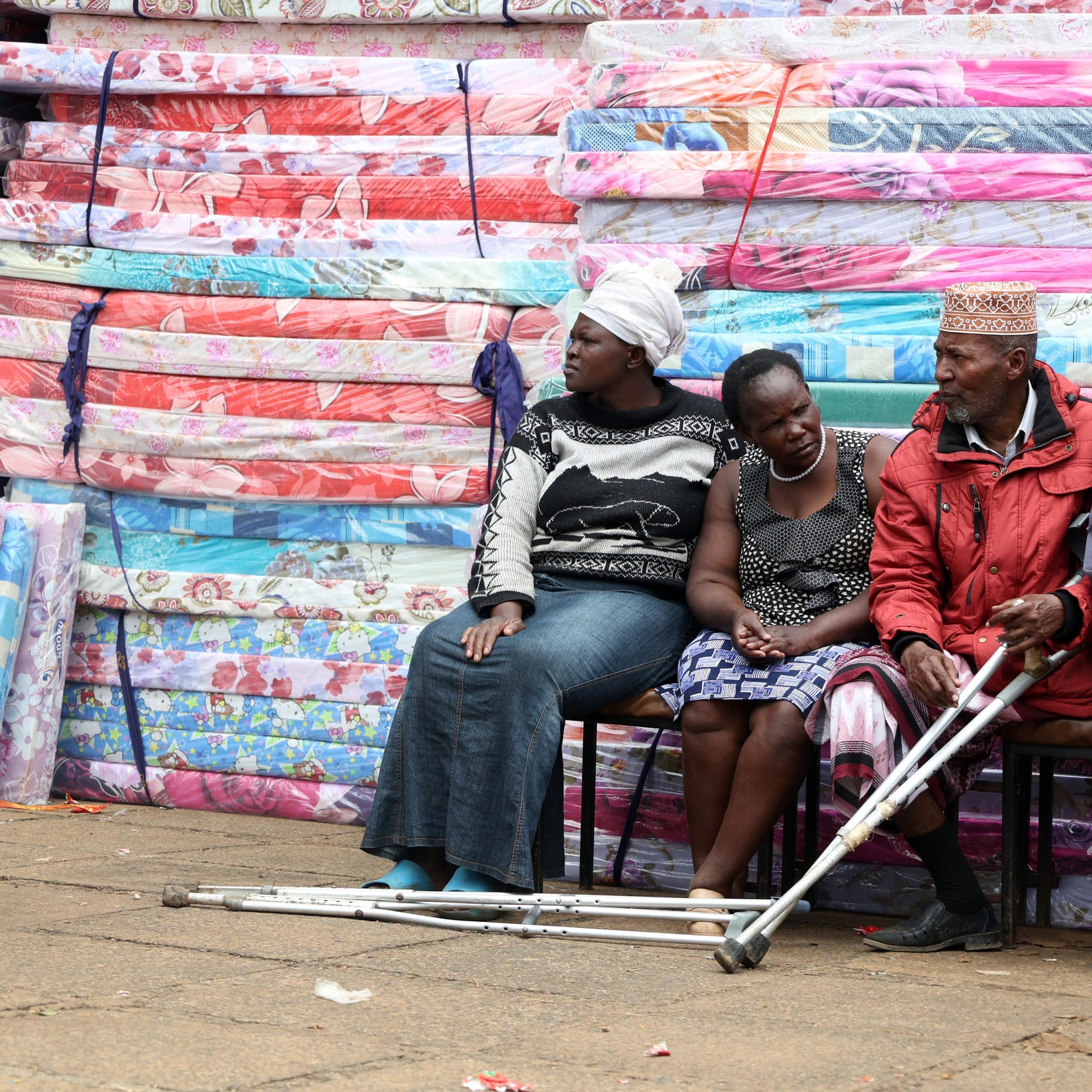 Four people, one of them with crutches, seated next to a pile of mattresses wrapped in plastic