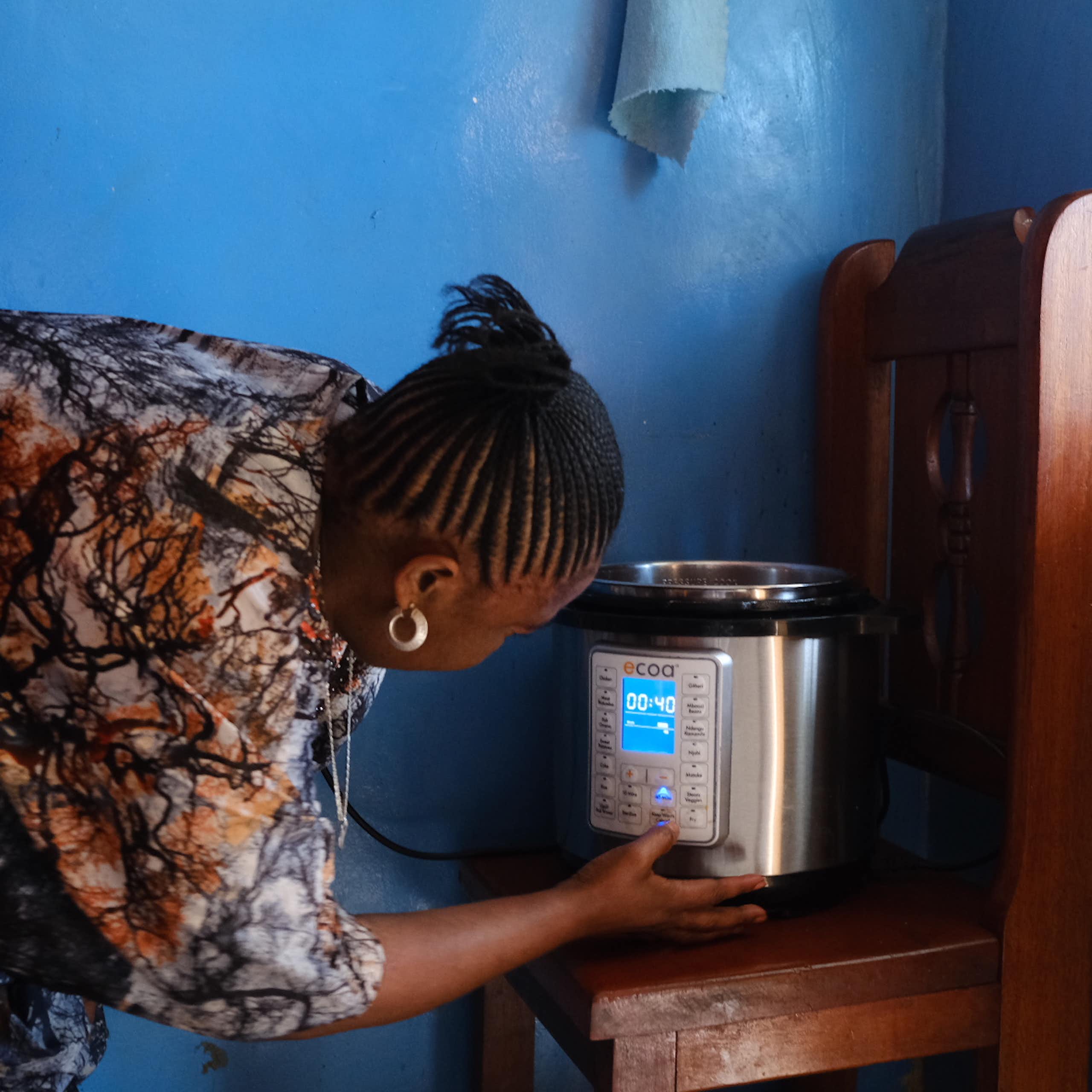 A woman turning on an electric pressure cooker that is standing on a wooden chair with a bright blue wall in the background
