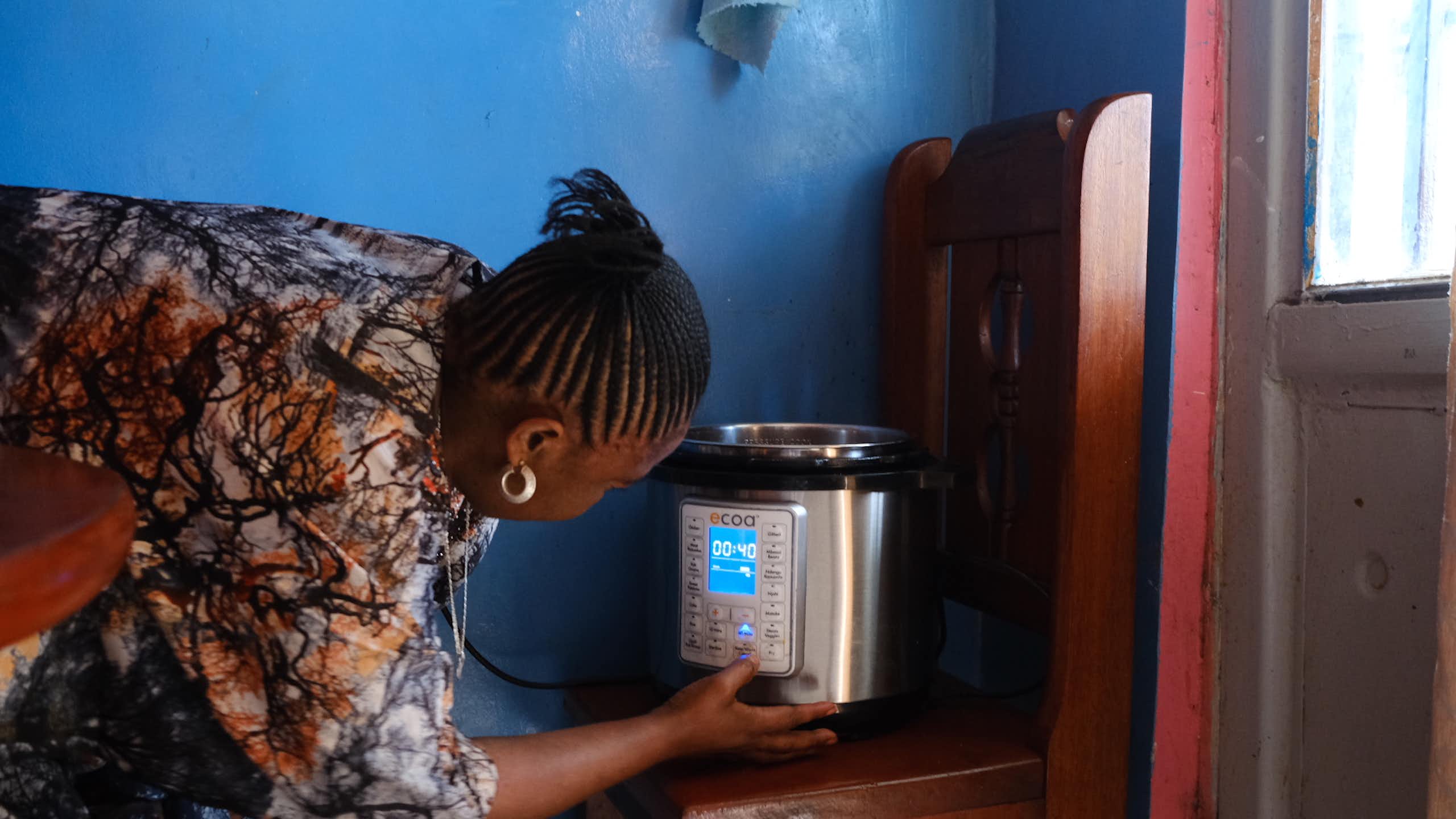 A woman turning on an electric pressure cooker that is standing on a wooden chair with a bright blue wall in the background