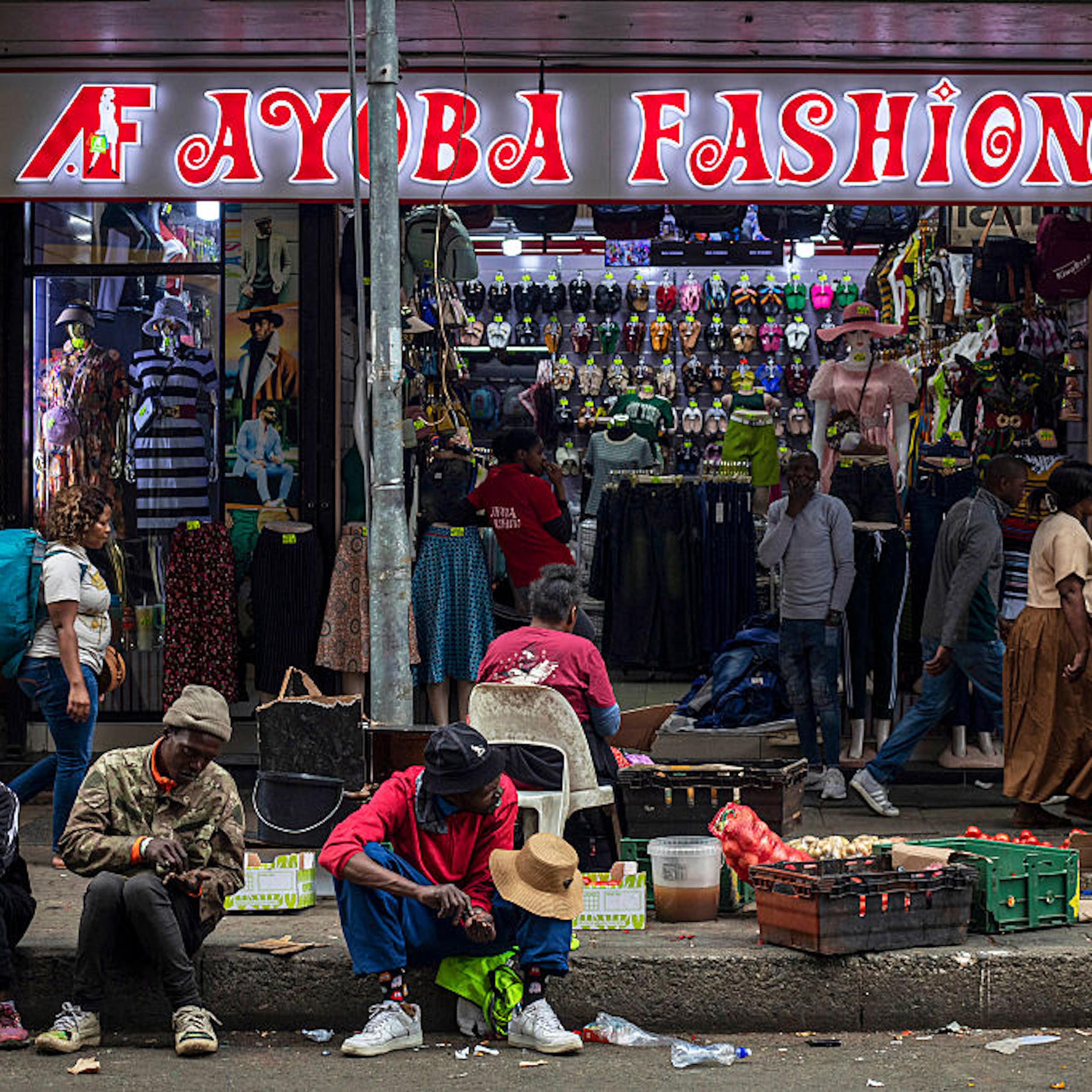 A small clothing shop and people selling vegetables on the pavement outside. Pedestrians walk past. Some men are sitting on the pavement facing the road.