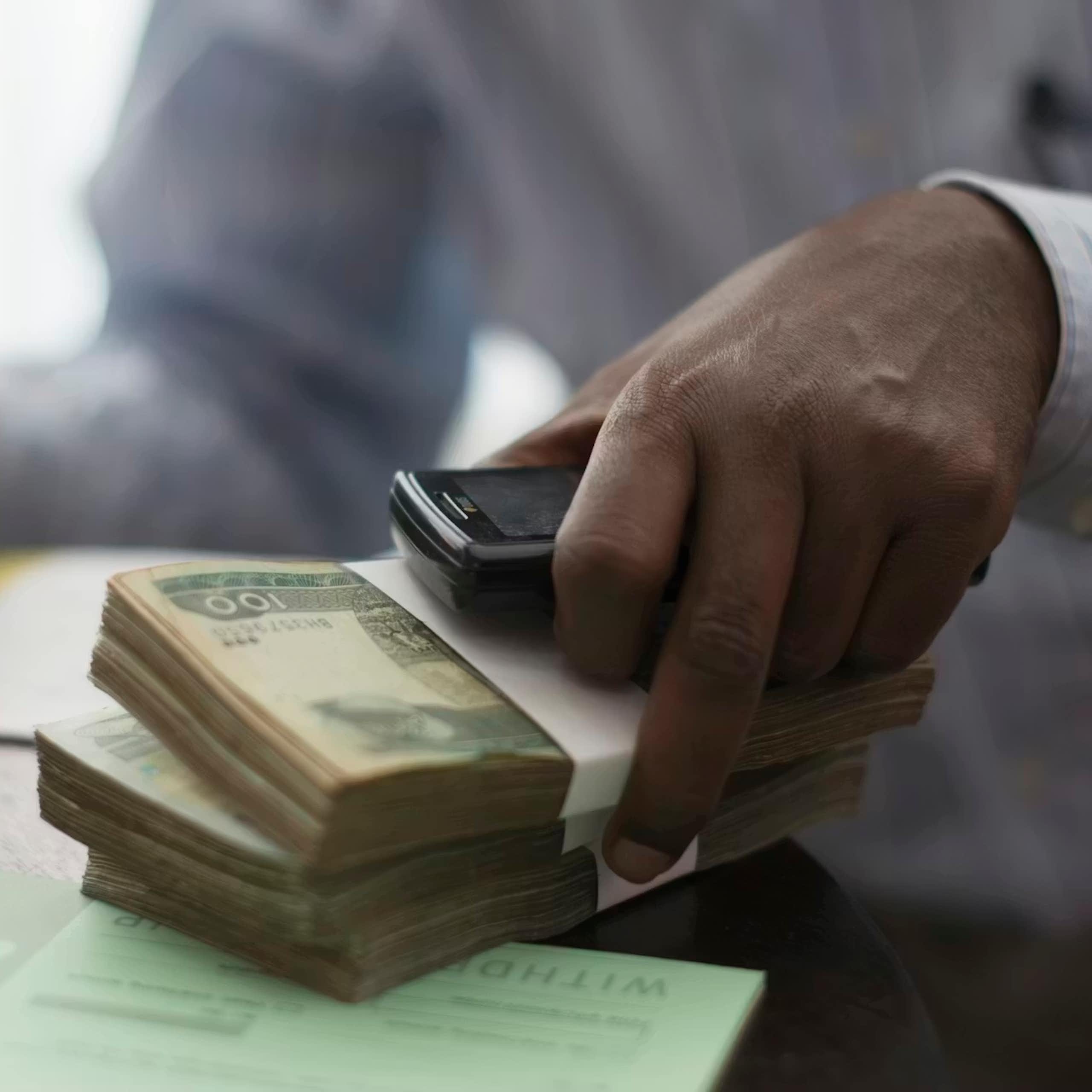 An African man's hands working at a desk, with two piles of cash in one hand he writes on a notepad with the other.