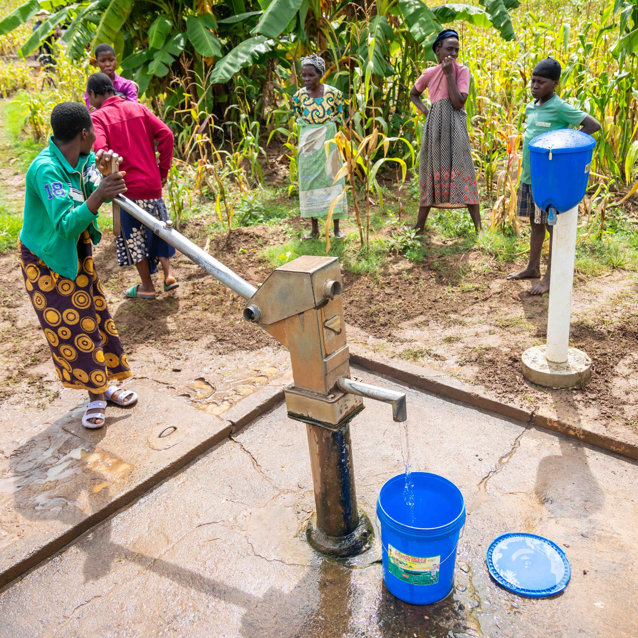 Five people stand with buckets near a tall water pump that is operated by pumping it by hand.