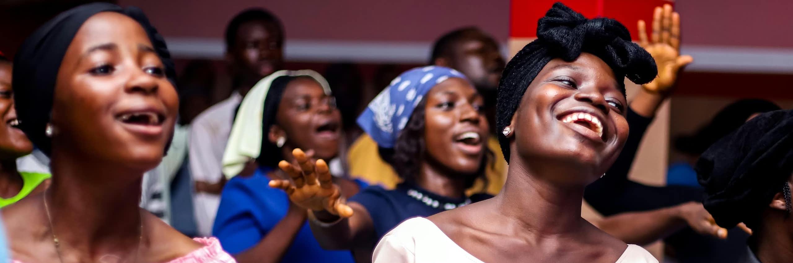 Two young African women smile as they sing, surrounded by other people in a gathering, some with raised arms.