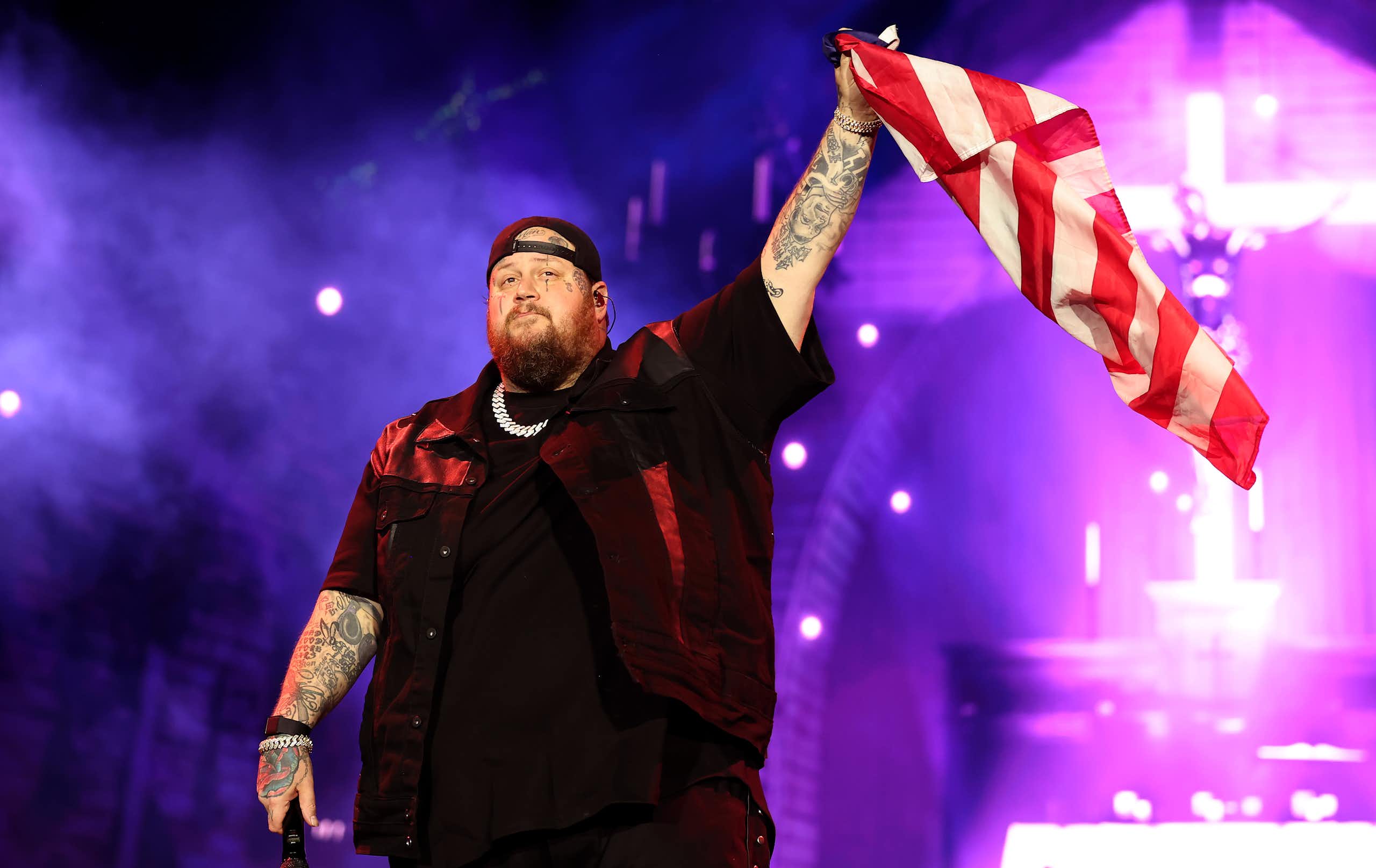 A heavily-tattooed singer in black clothes waves the US flag onstage at a concert.