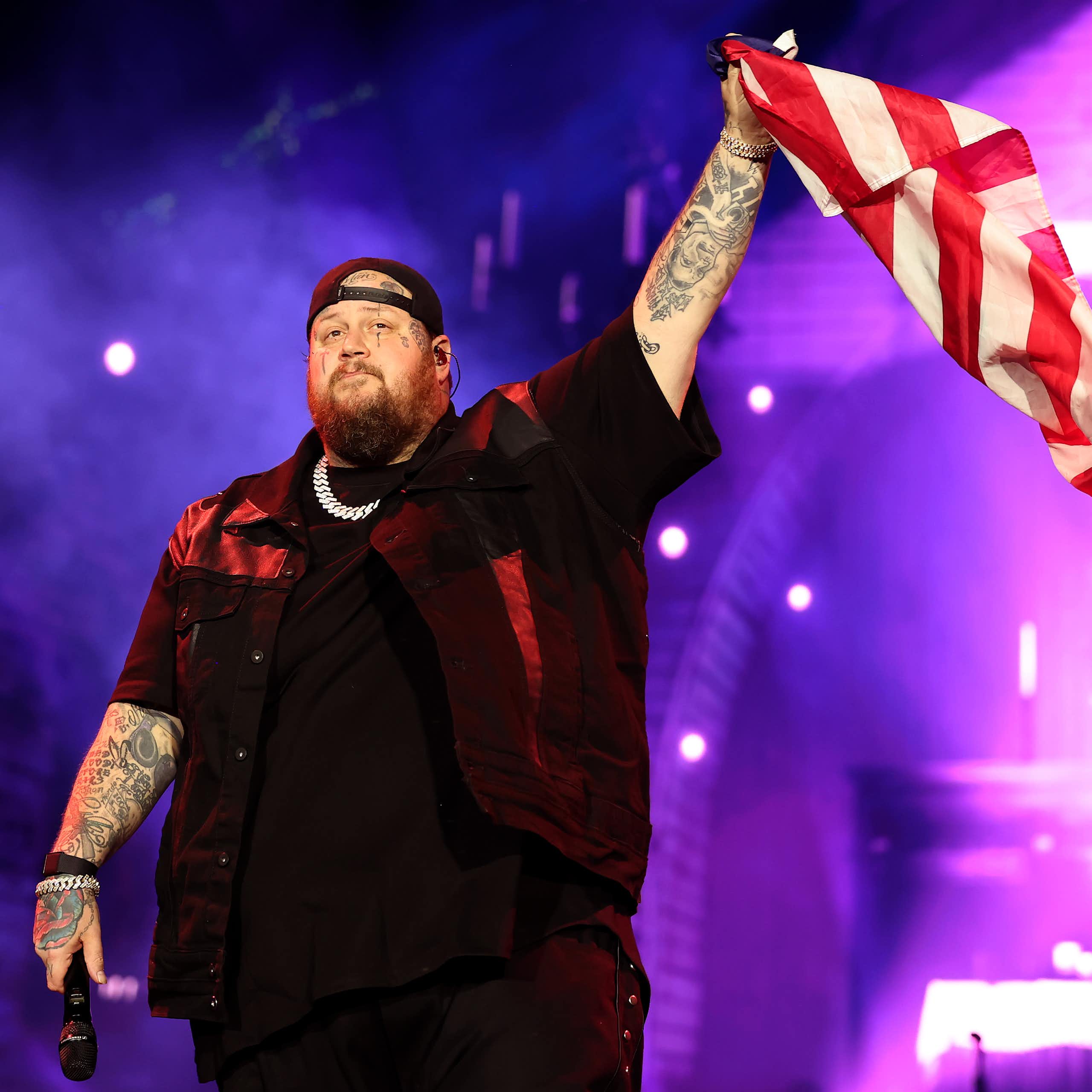 A heavily-tattooed singer in black clothes waves the US flag onstage at a concert.