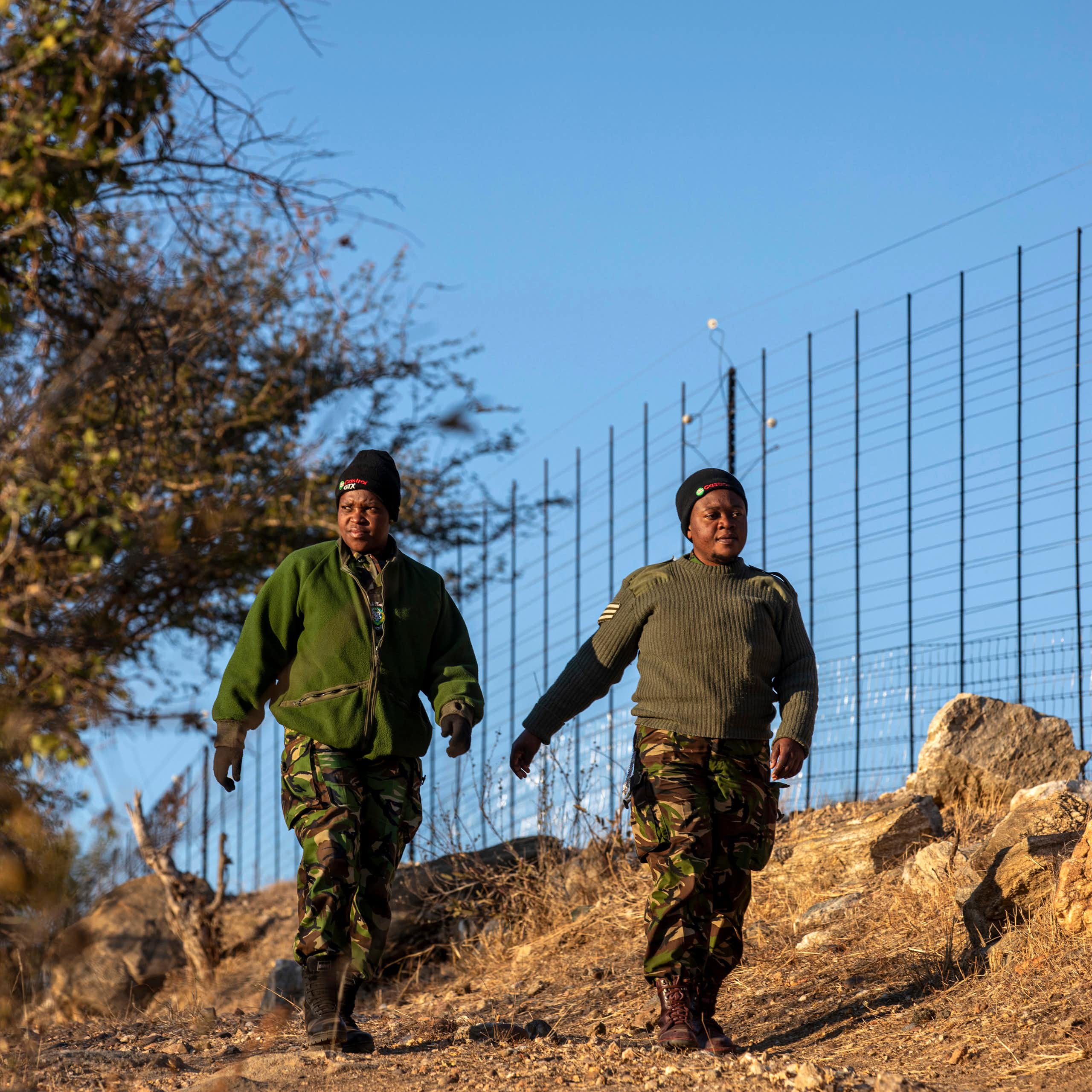 Two women in green uniforms walk next to a tall fence
