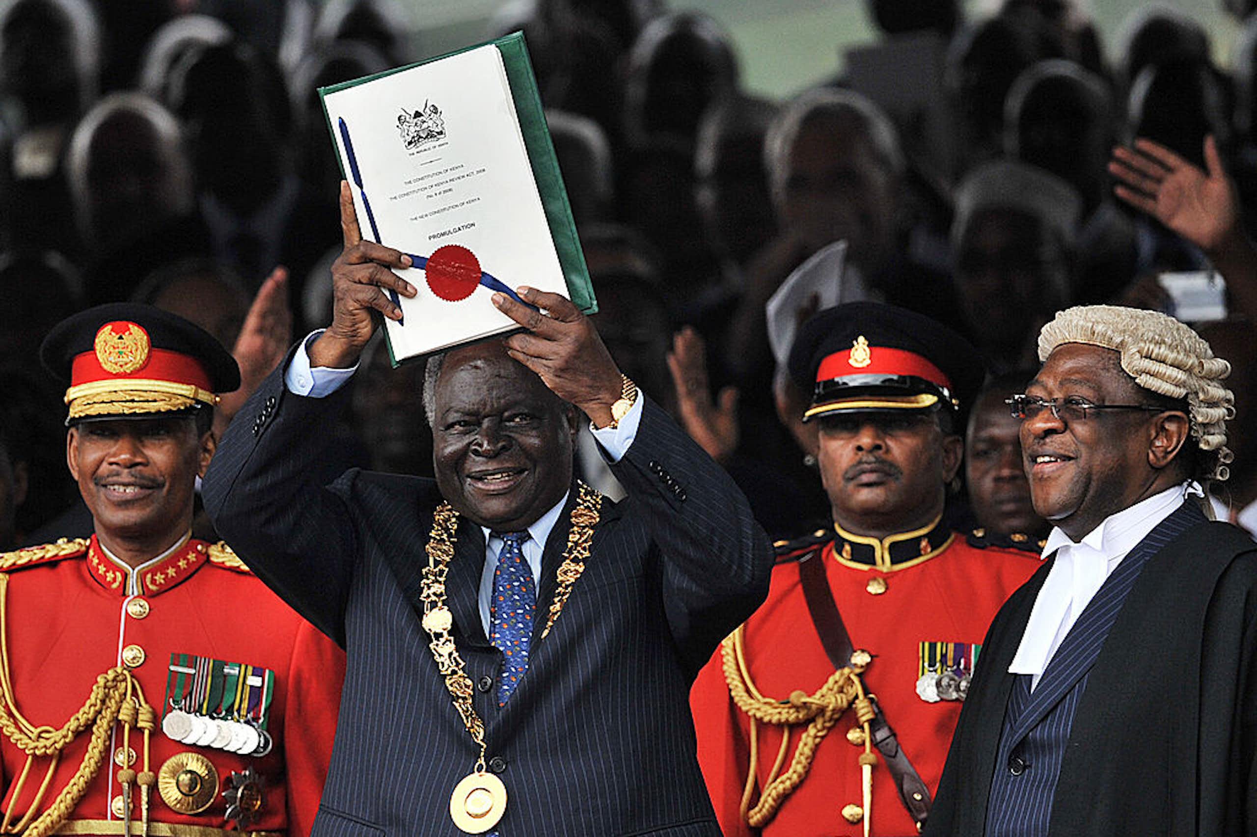 Man dressed in a suit with a gold mace around his neck holding up a document while other people around him smile