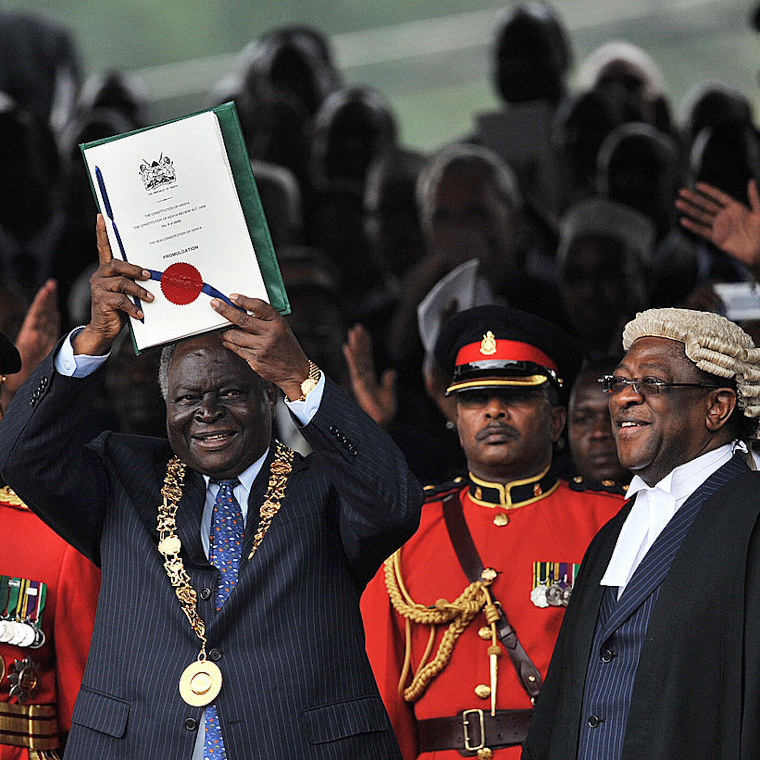 Man dressed in a suit with a gold mace around his neck holding up a document while other people around him smile