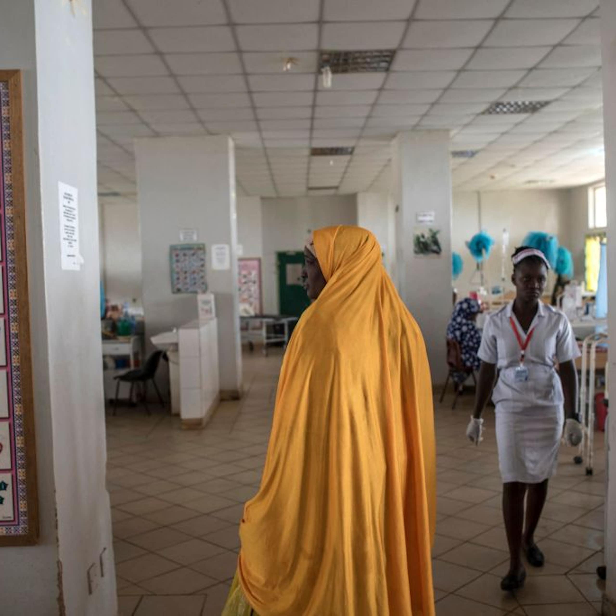 Woman in a robe stands in a hospital ward as a nurse approaches