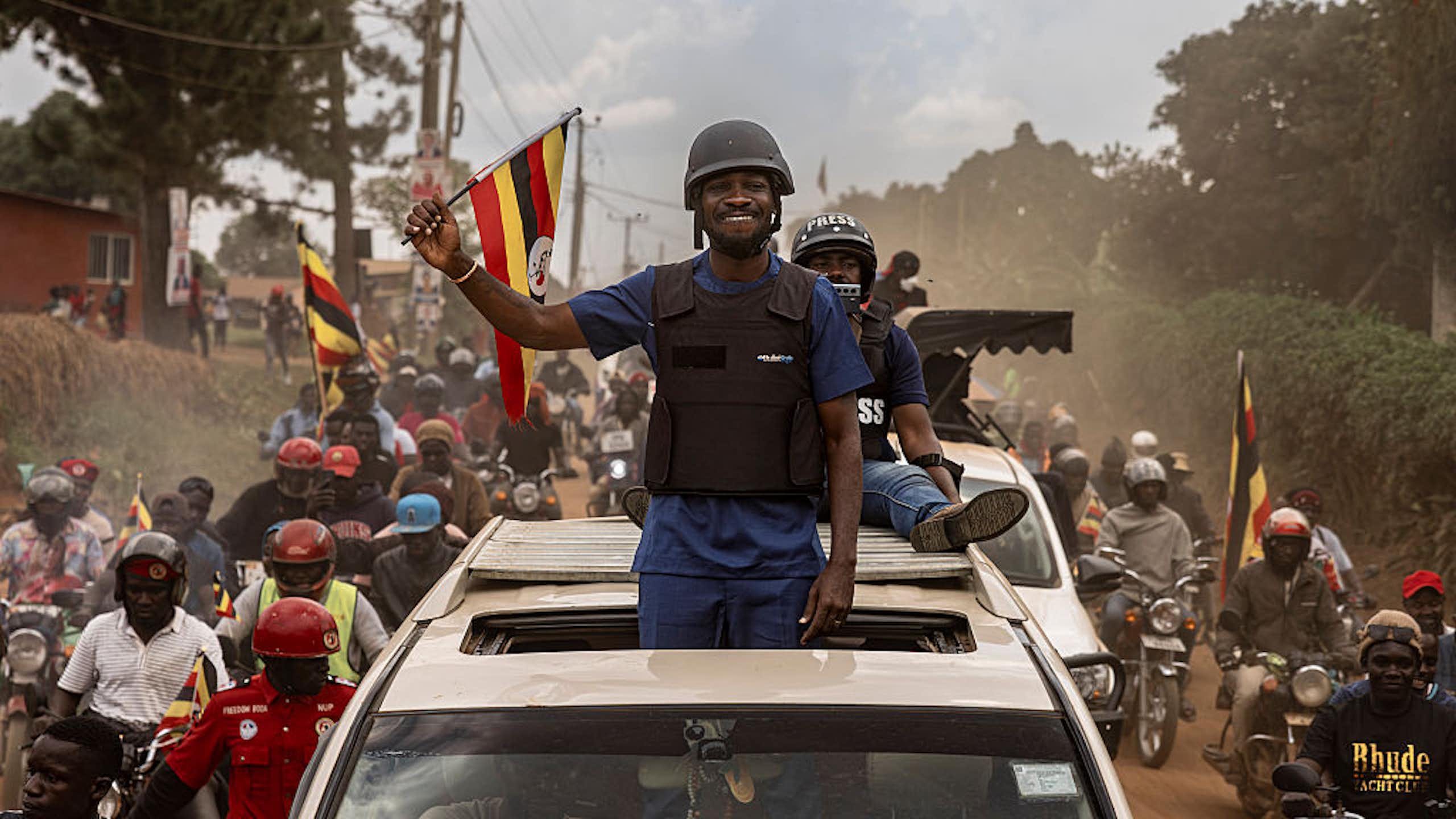A man wearing a bulletproof vest standing from the sunroof of a vehicle and waving to a crowd of people, some of whom are waving flags.