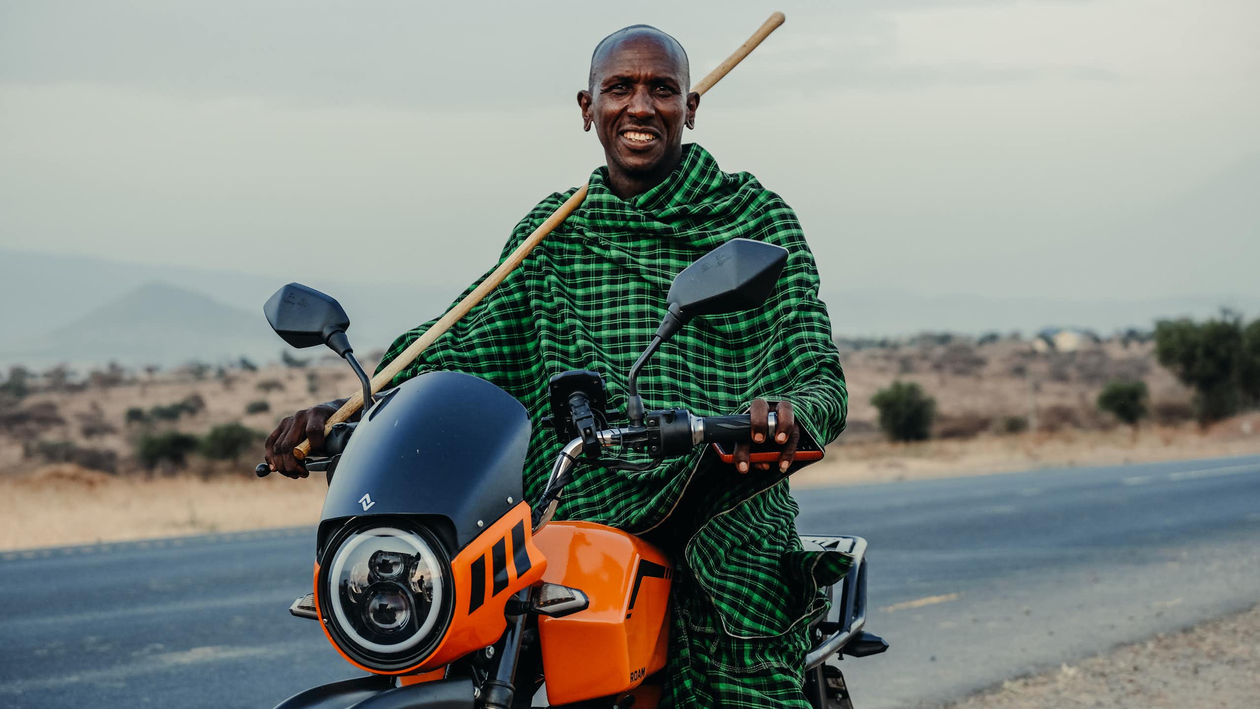 Man in checked shawl, holding a stick and sitting on a motorbike