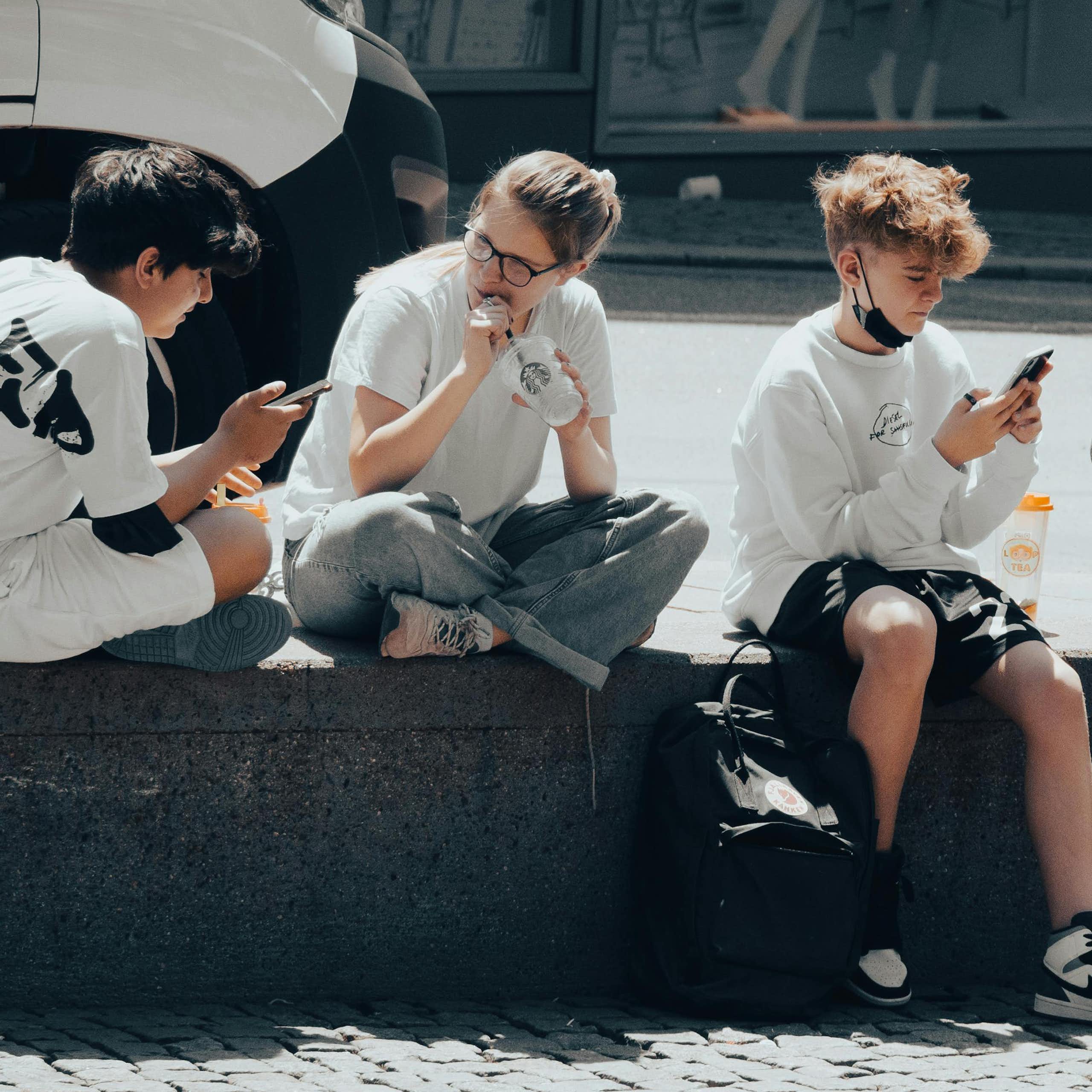 A group of teenagers sitting on the street, looking at their phones and talking. 