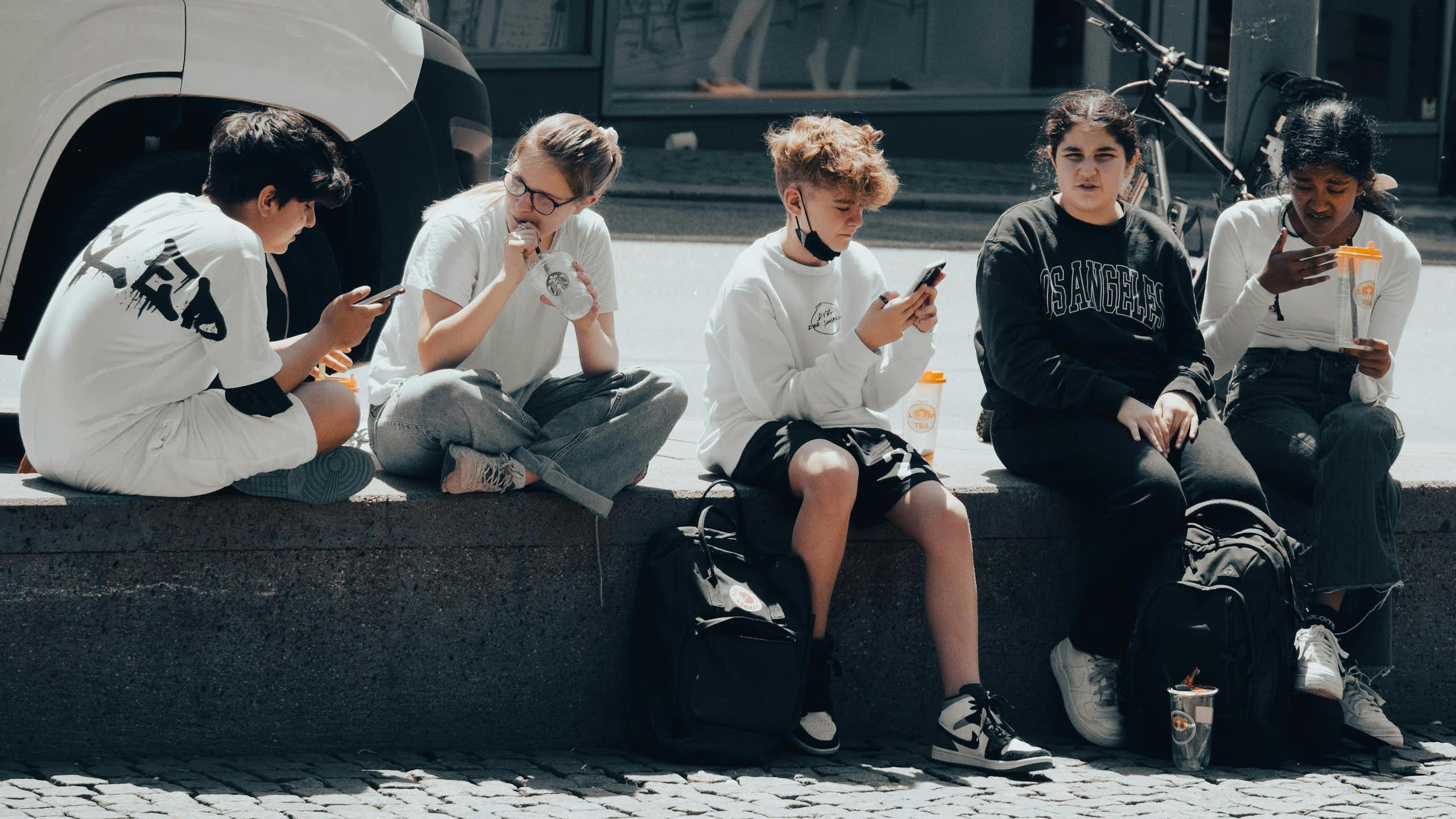 A group of teenagers sitting on the street, looking at their phones and talking. 