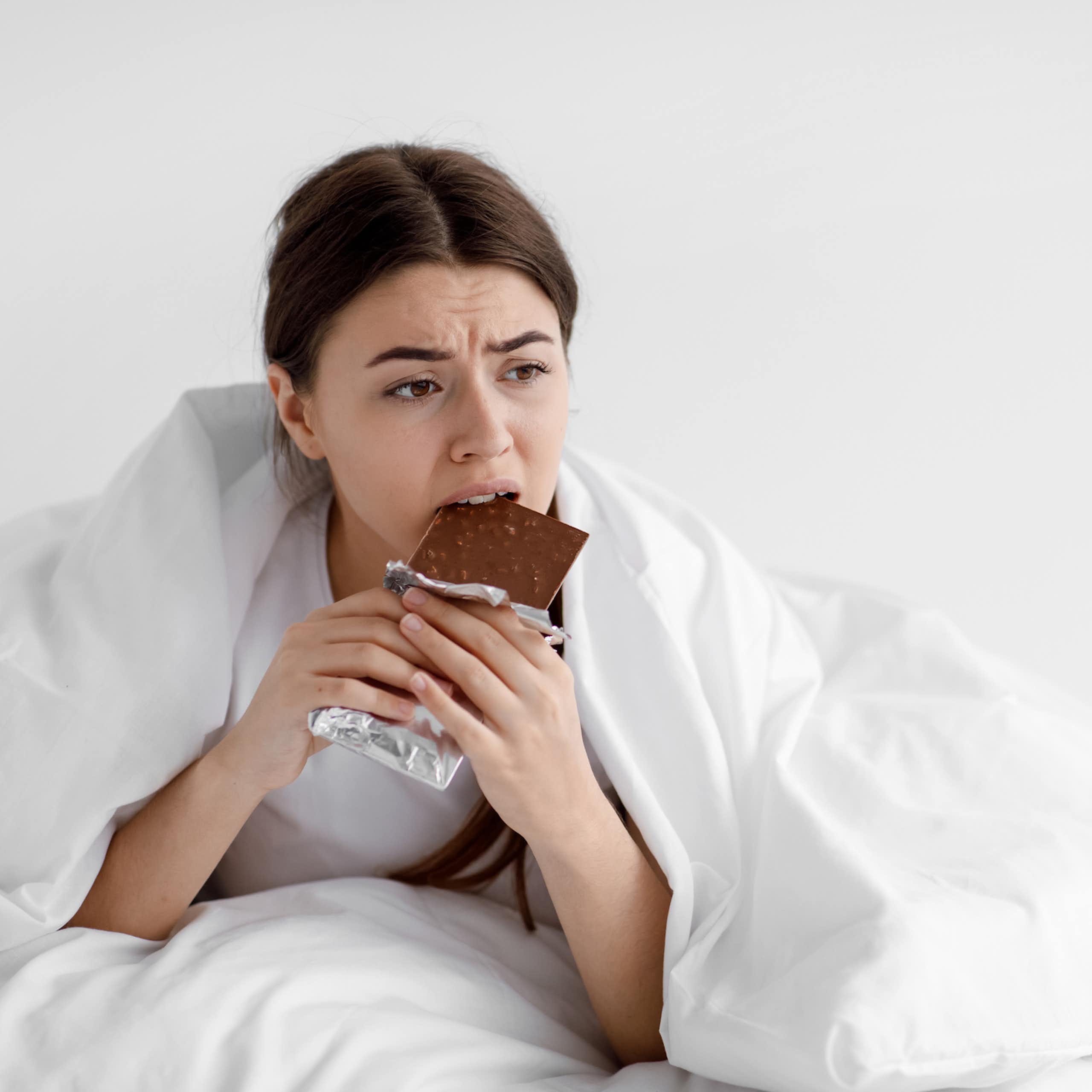 Woman eats chocolate while wrapped in bedsheets.