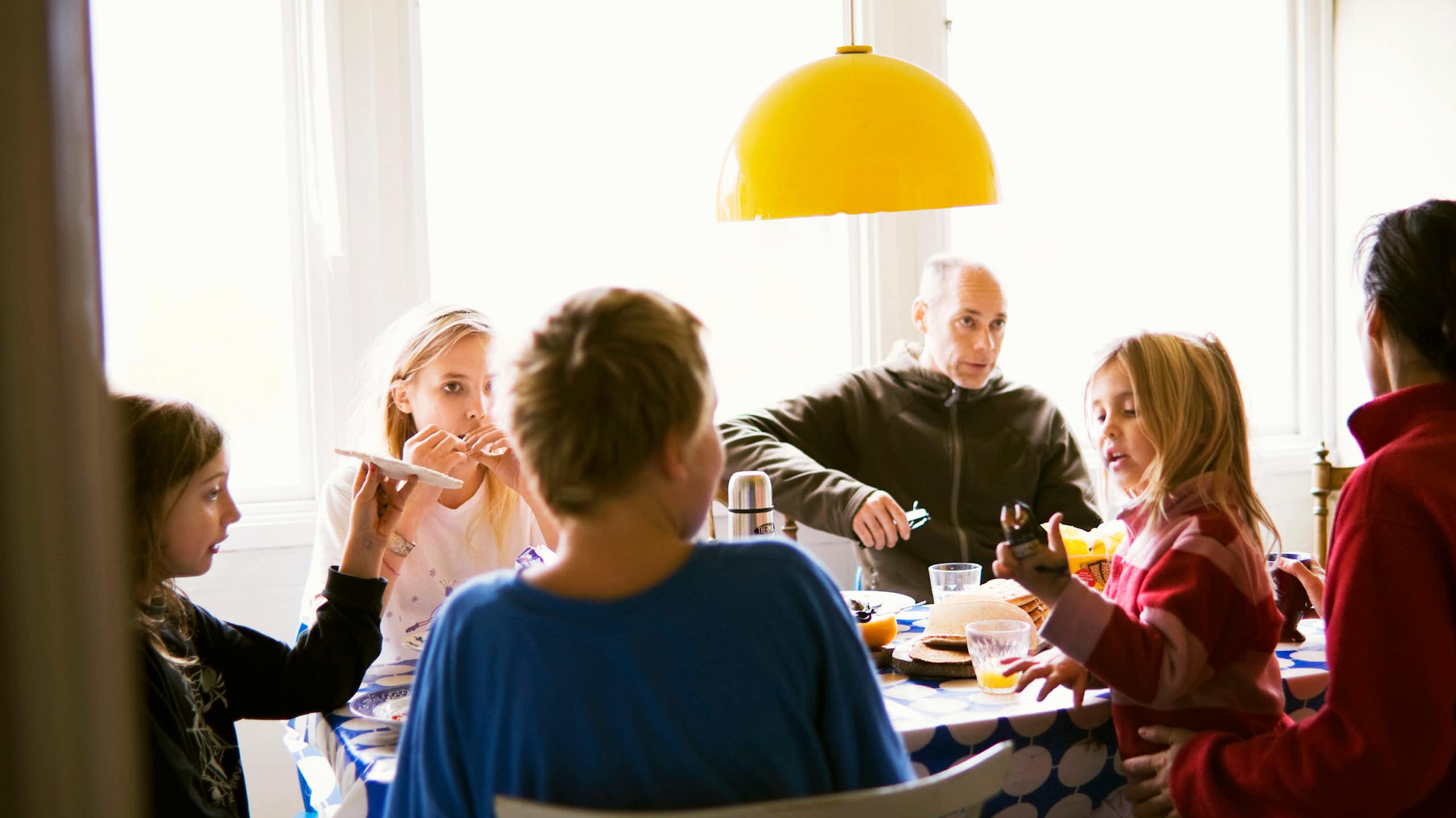A family group sits at a table having a meal. A young child stands a man wears a serious expression. 