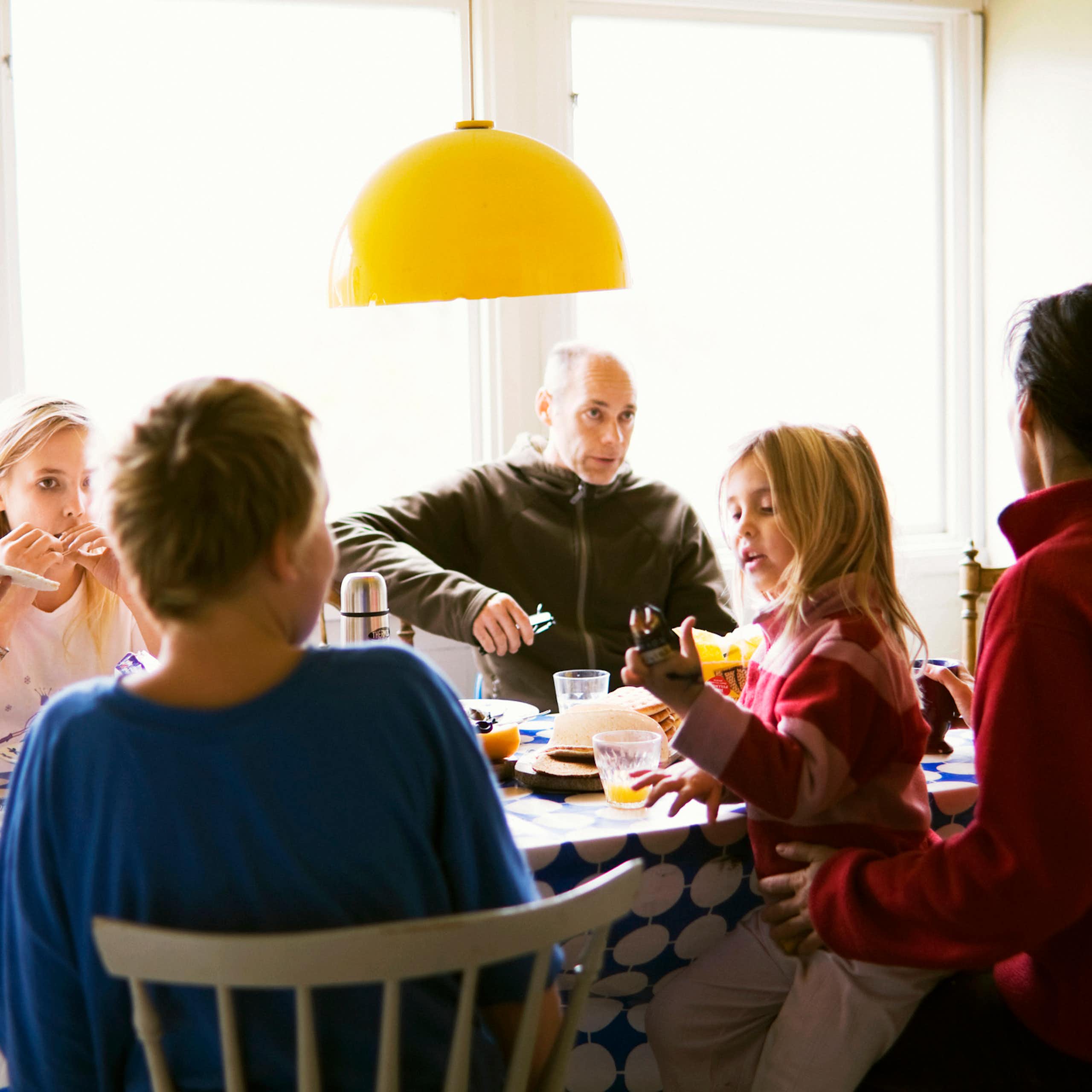 A family group sits at a table having a meal. A young child stands a man wears a serious expression. 