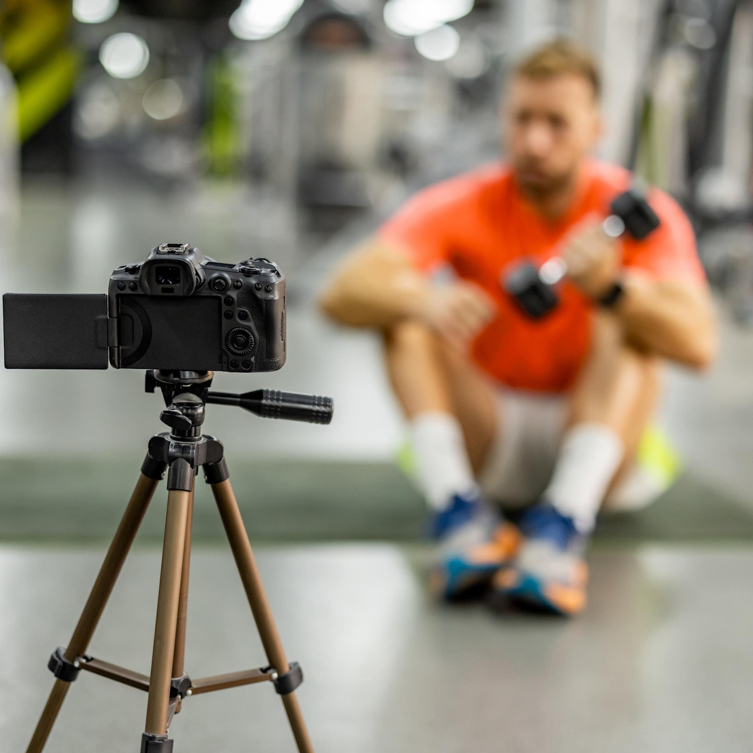 A blurry image of a man in an orange shirt lifting a weight as he sits on the floor in front of a camera tripod.
