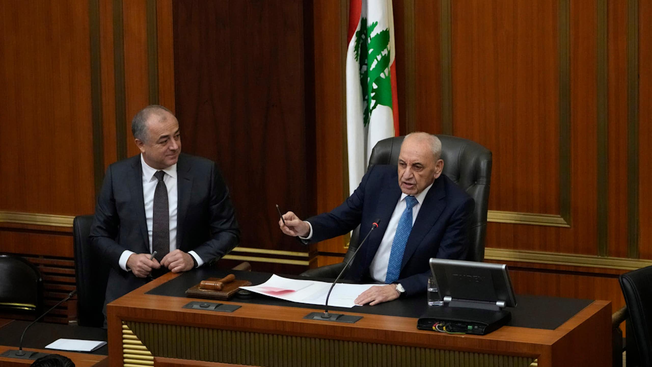 A men in a suit gestures from his speaker's chair.