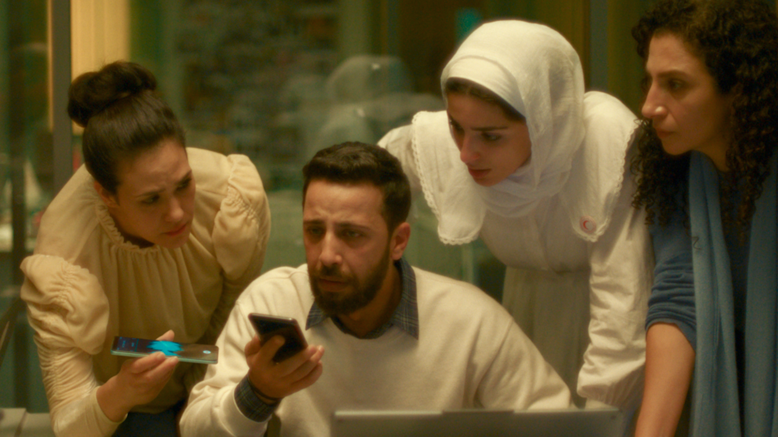 Three women, one in a headscarf, stand anxiously above a man seated with a phone in his hand.