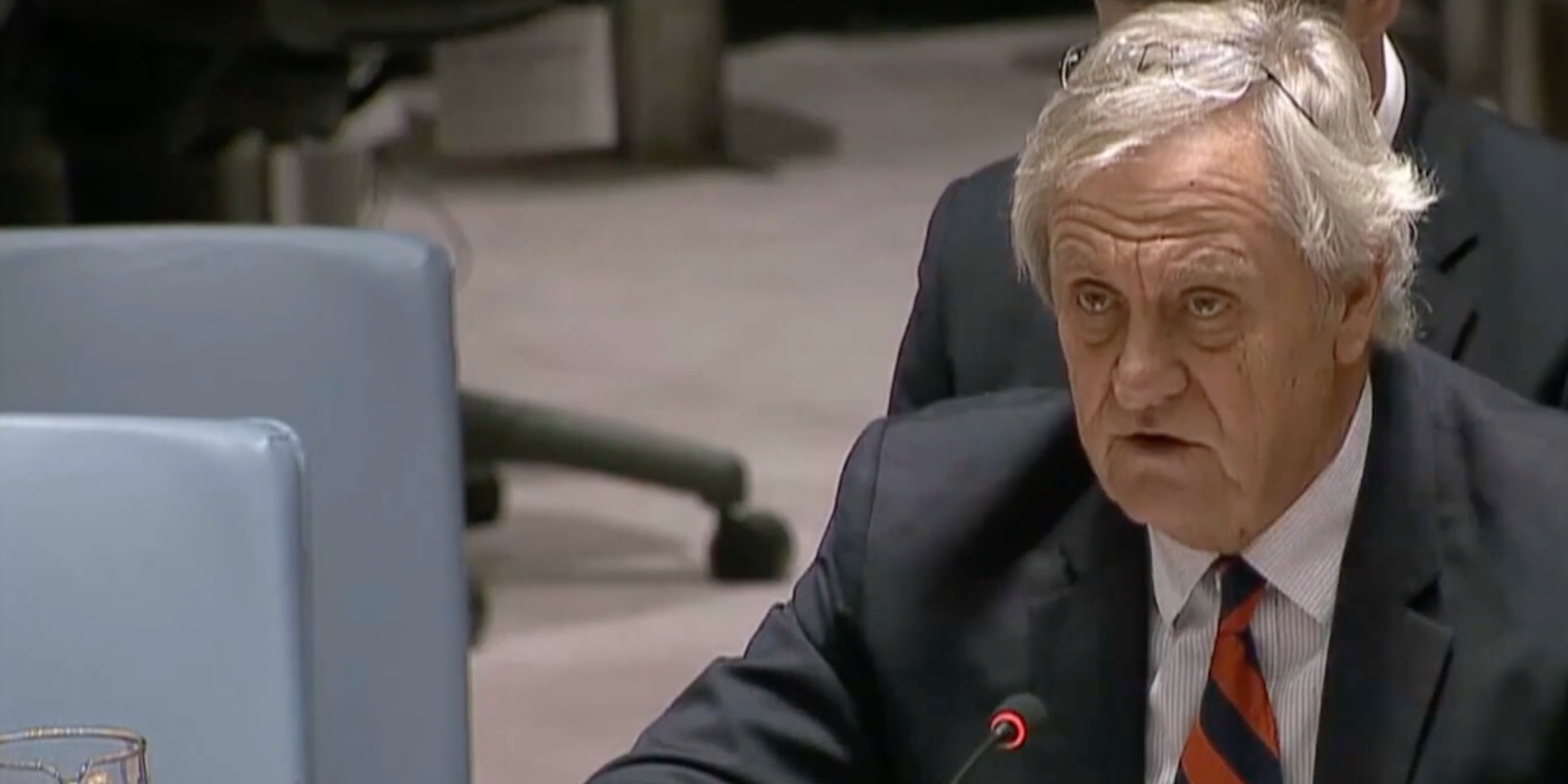 A man in a suit and tie, seated at a microphone