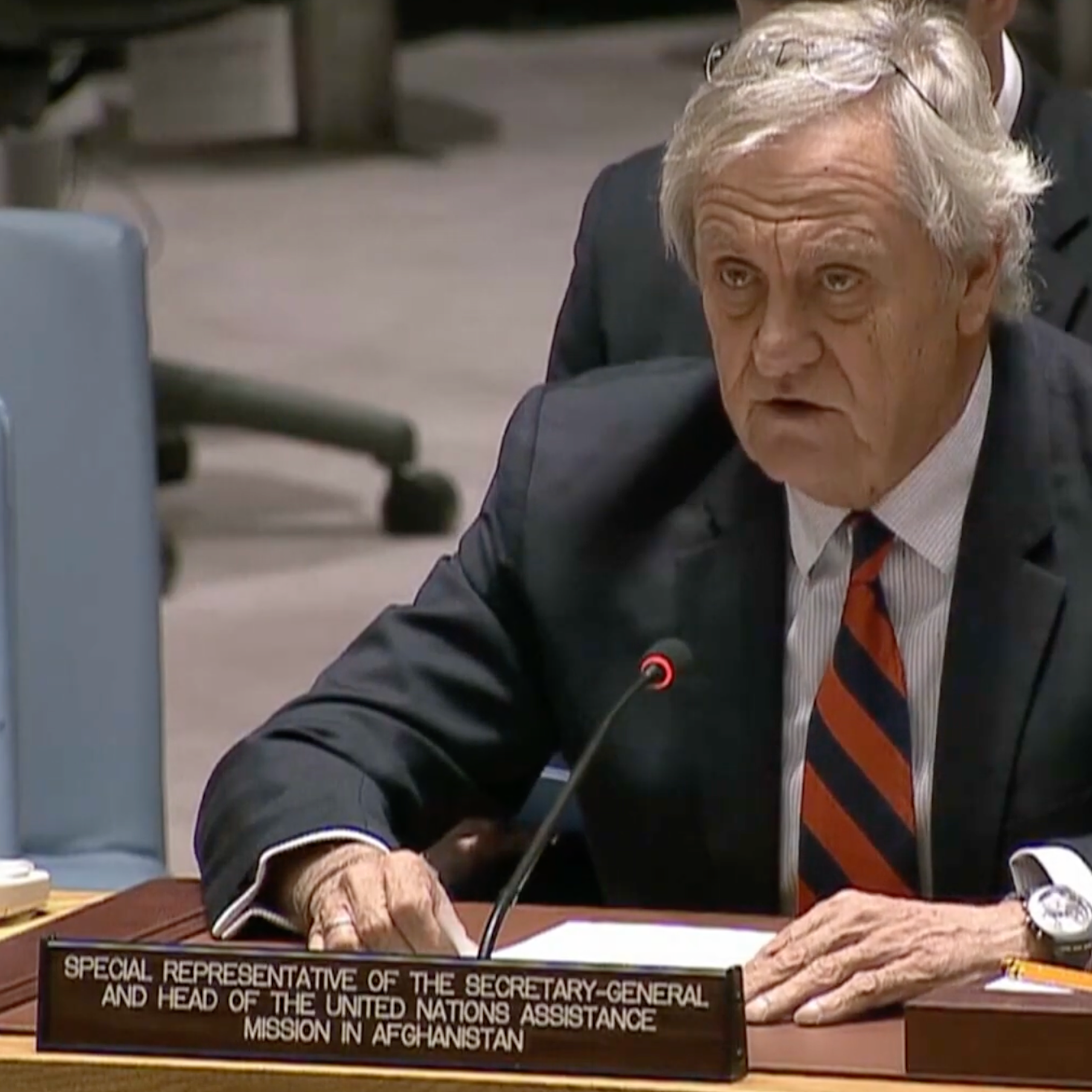 A man in a suit and tie, seated at a microphone